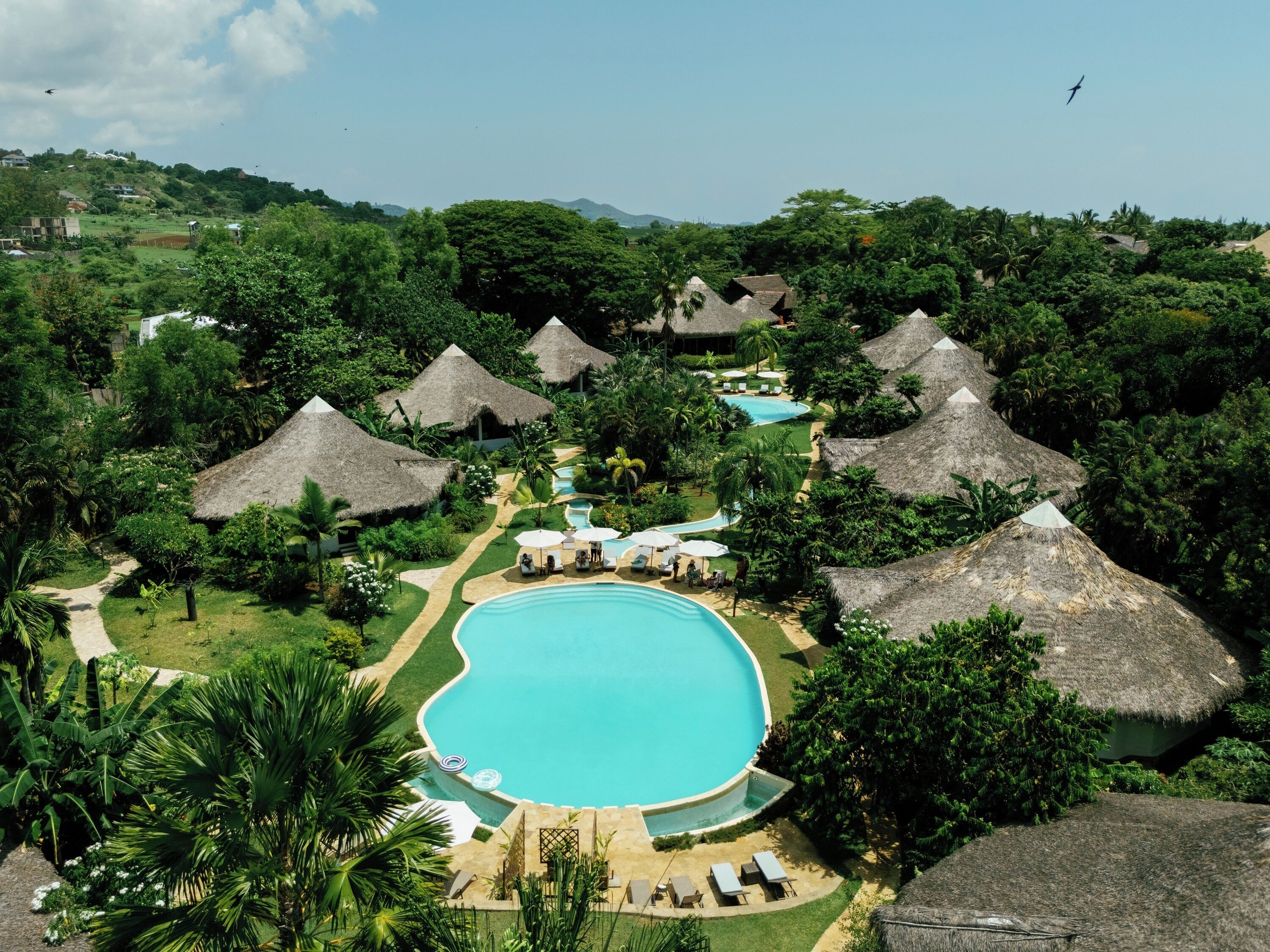 A resort with a central swimming pool, thatched-roof huts, and lush greenery.