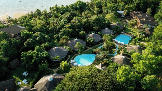 An aerial view of a resort with multiple swimming pools surrounded by lush greenery and thatched-roof structures. - Loharano Lodge (Nosy Be)