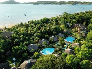 An aerial view of a resort with multiple swimming pools surrounded by lush greenery and thatched-roof structures. - Loharano Lodge (Nosy Be)