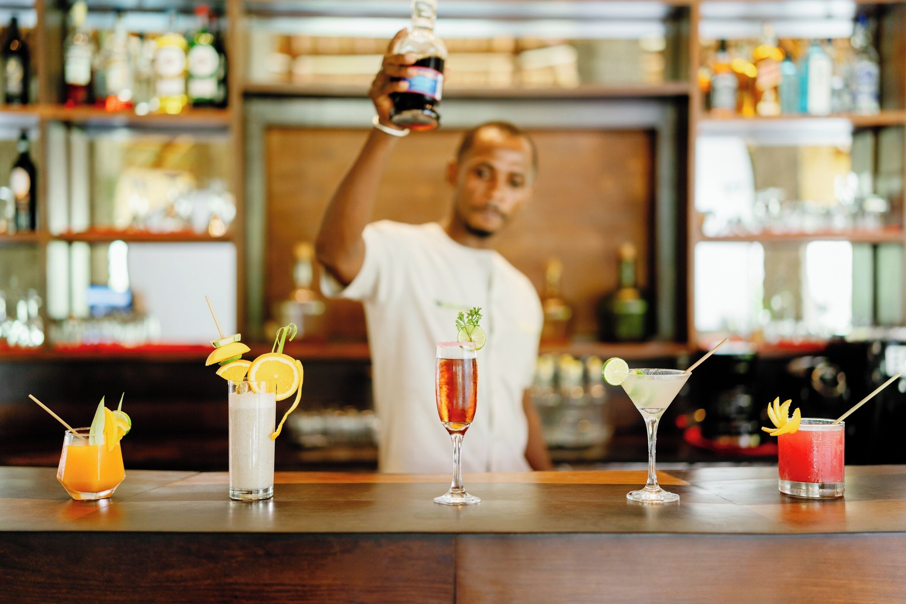 A bar with various cocktails on the counter, a bartender preparing drinks, and shelves stocked with bottles in the background.
