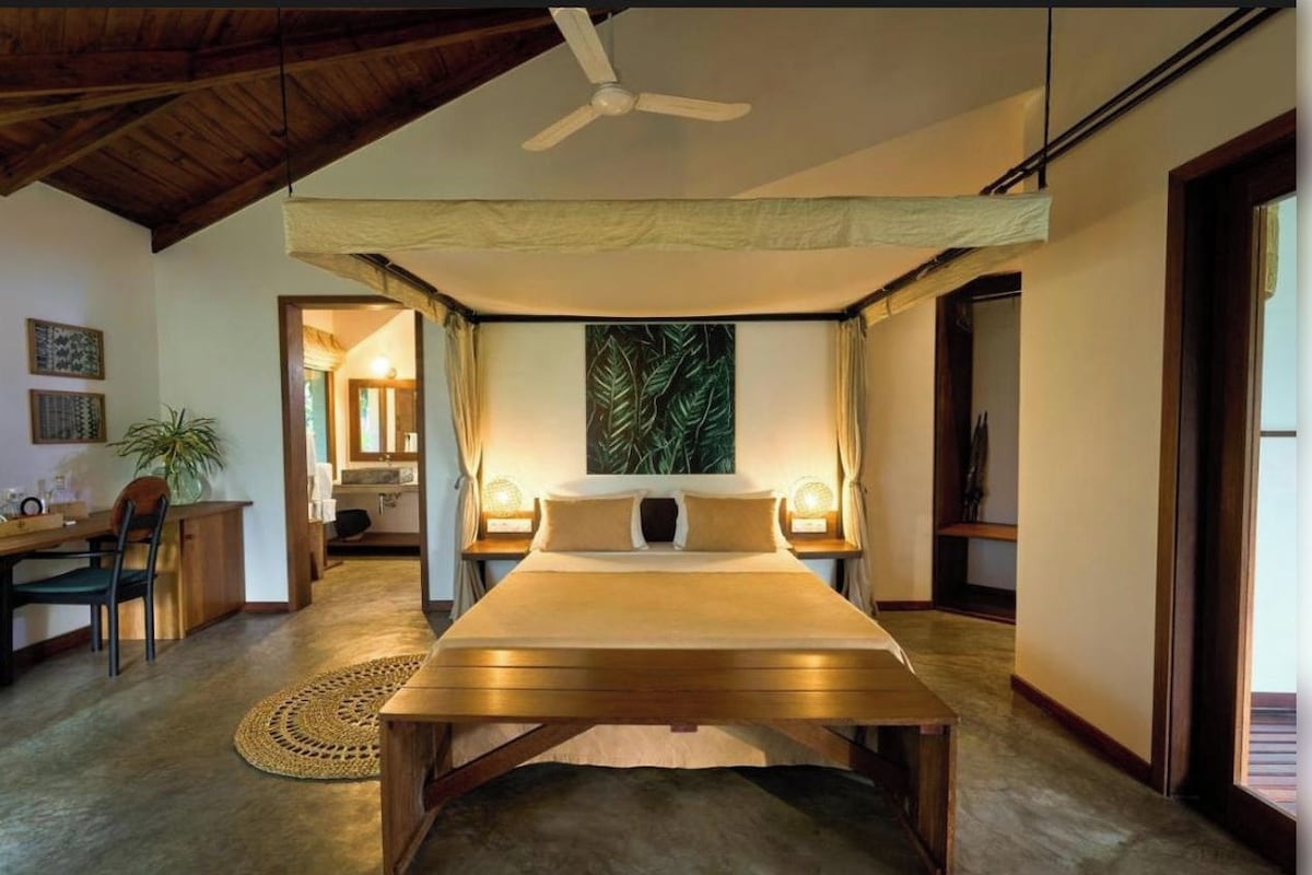 A bedroom with a bed, a ceiling fan, a large painting, and a desk with a chair.