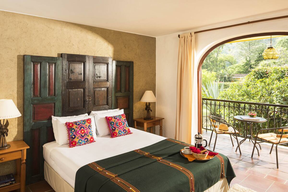 A hotel room with a large bed, a balcony with a table and chairs, and a view of greenery outside.