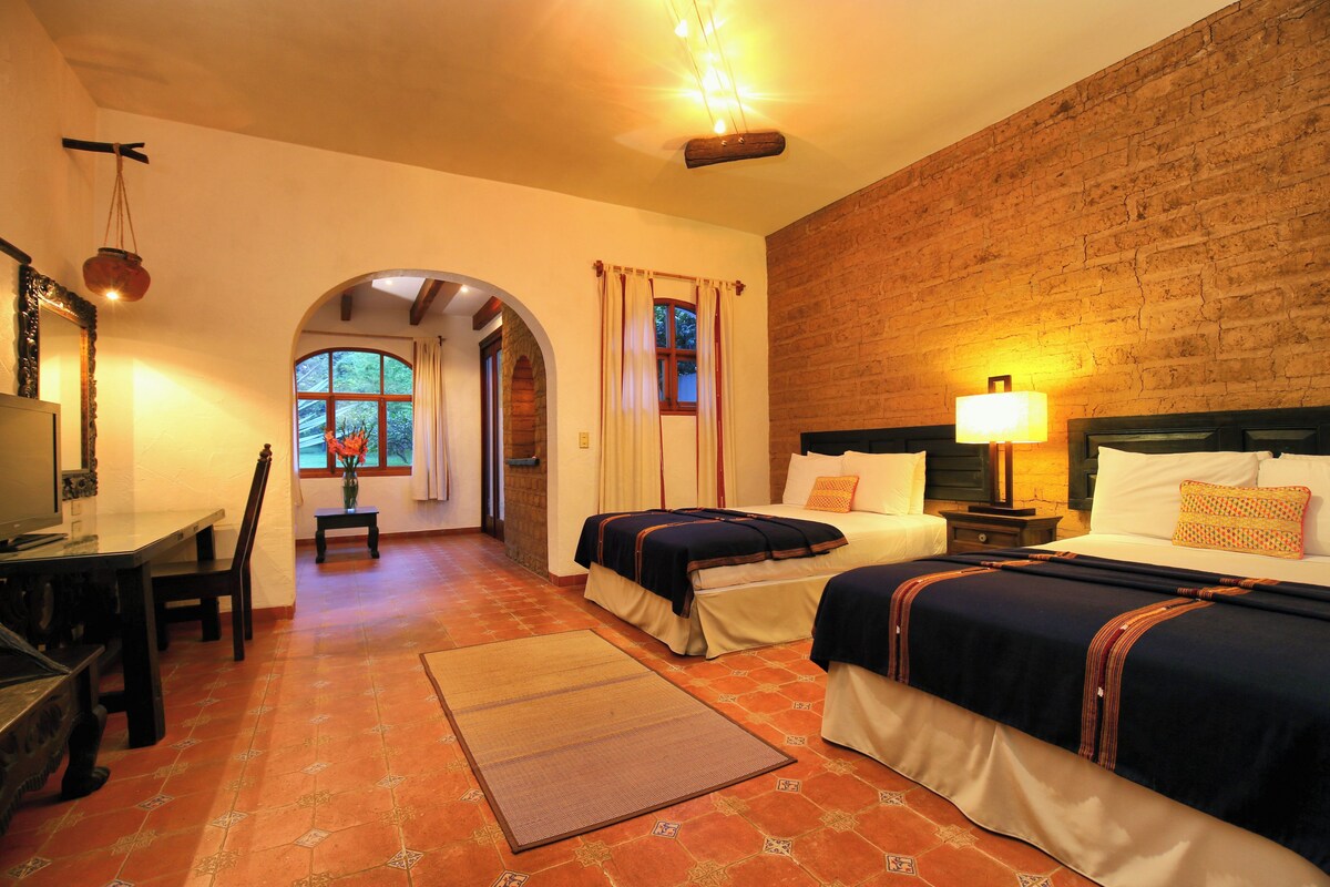 A hotel room with two beds, a desk, and a chair. There is a brick wall and a window with curtains.