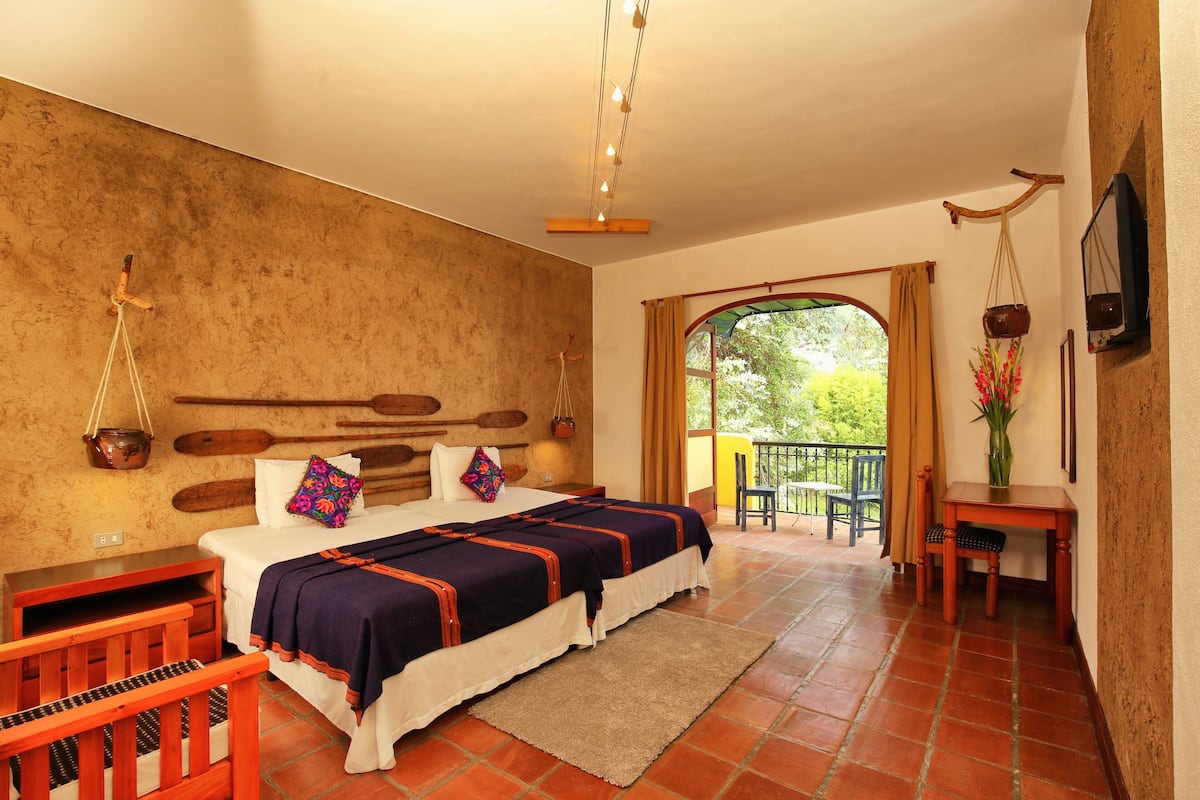 A bedroom with a large bed, wooden headboard, a small table with a chair, a TV, and a balcony with a view of greenery.