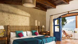 Une chambre spacieuse avec un grand lit, des poutres apparentes en bois au plafond et une vue sur la mer par une porte ouverte.