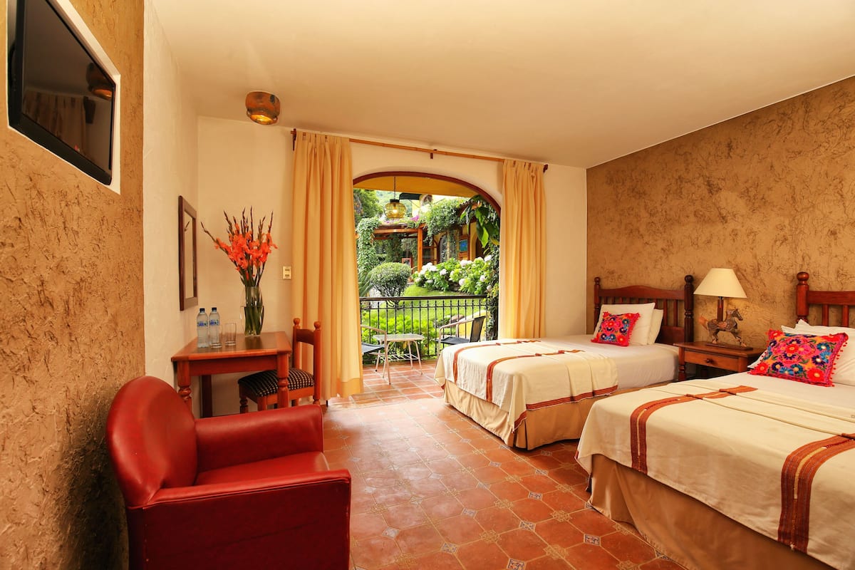 A hotel room with two beds, a red chair, a small table with a vase of flowers, and a view of a garden through an open door.