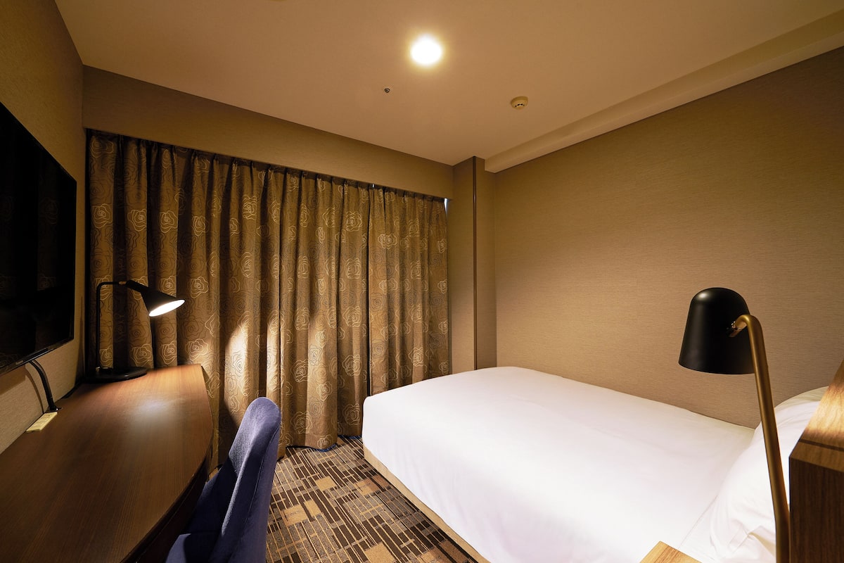 A hotel room with a bed, a desk with a lamp, a chair, and a TV.