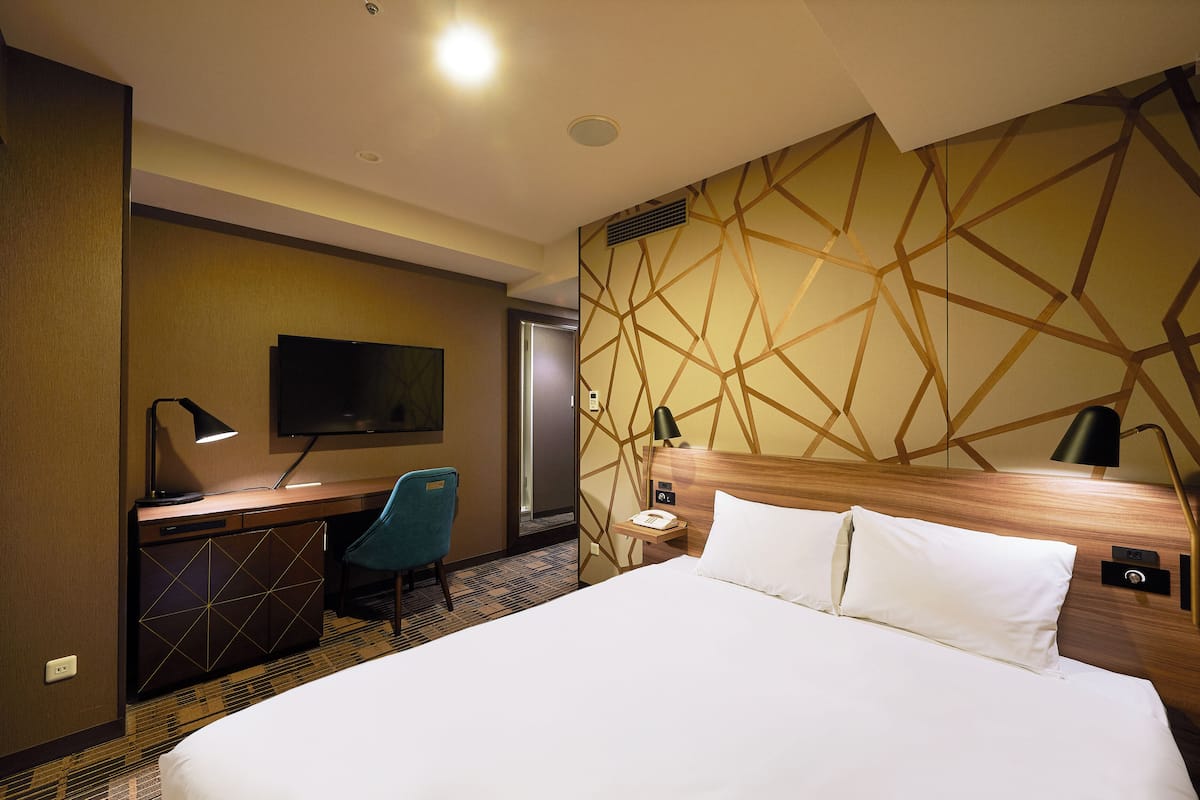 A hotel room with a large bed, a desk with a chair, a television, and a wall with a geometric pattern.