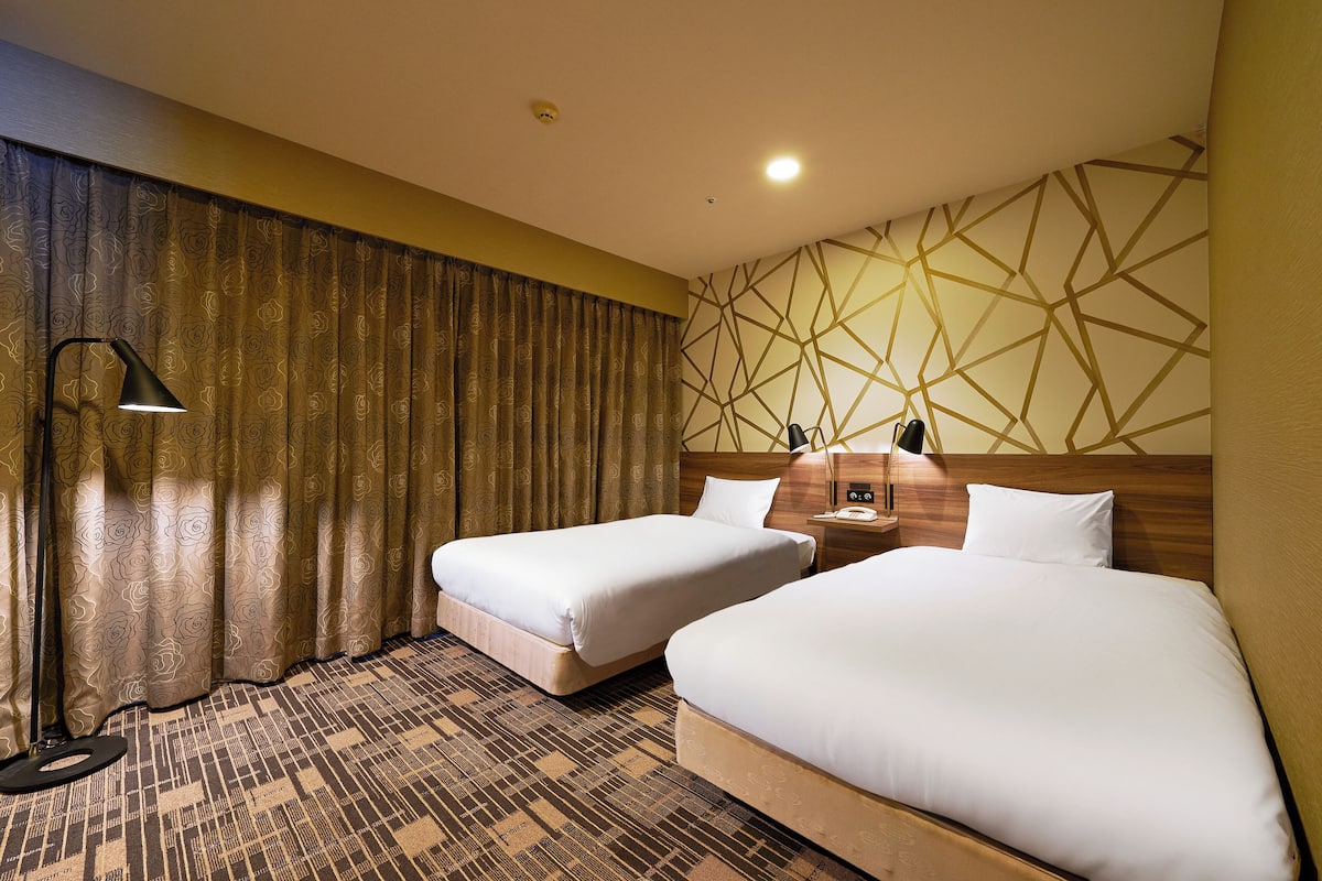 A hotel room with two beds, a patterned carpet, a wall with geometric design, and a lamp.