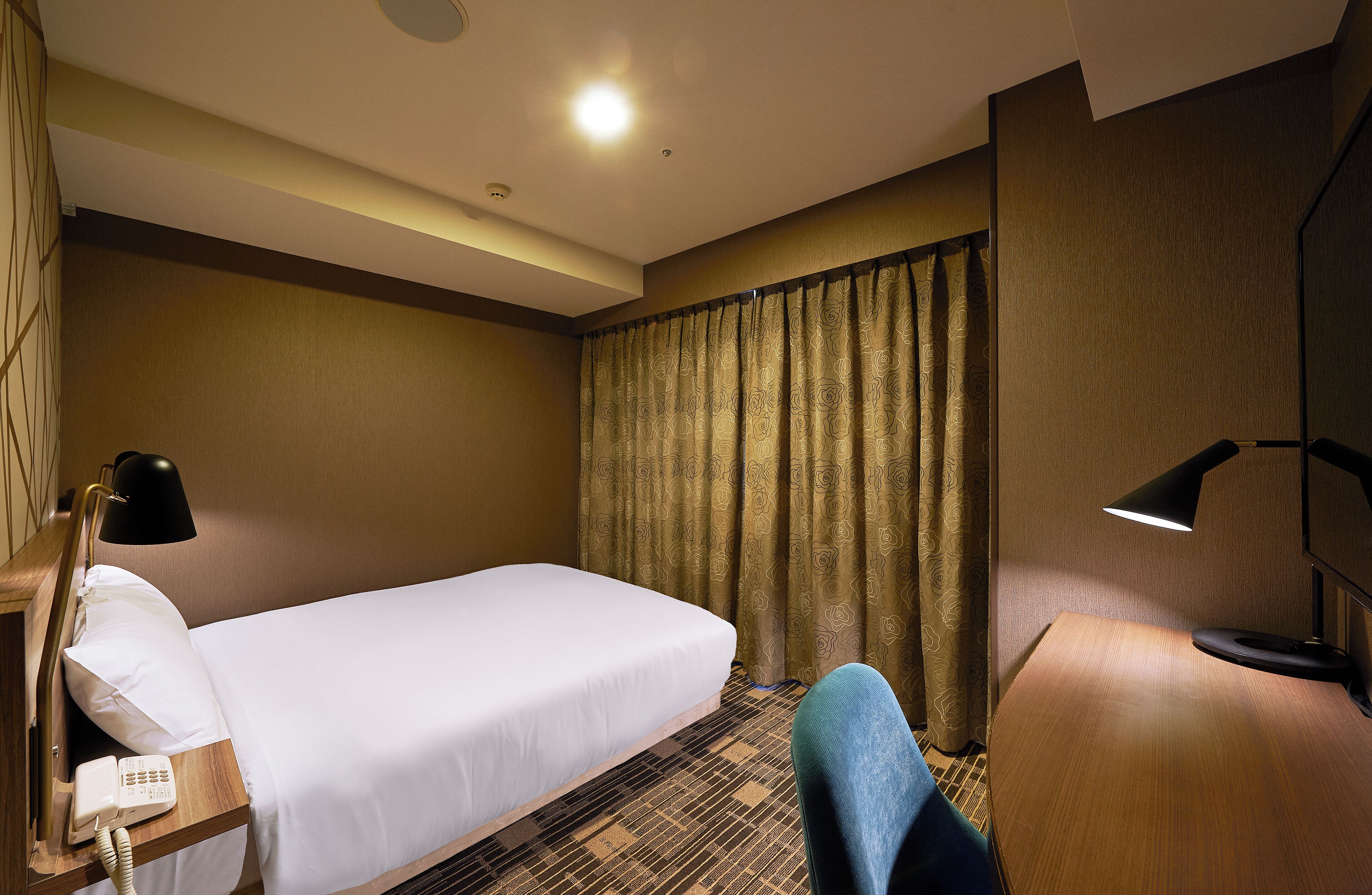 Una habitación de hotel con una cama individual, un escritorio con lámpara, un teléfono y una cortina.
