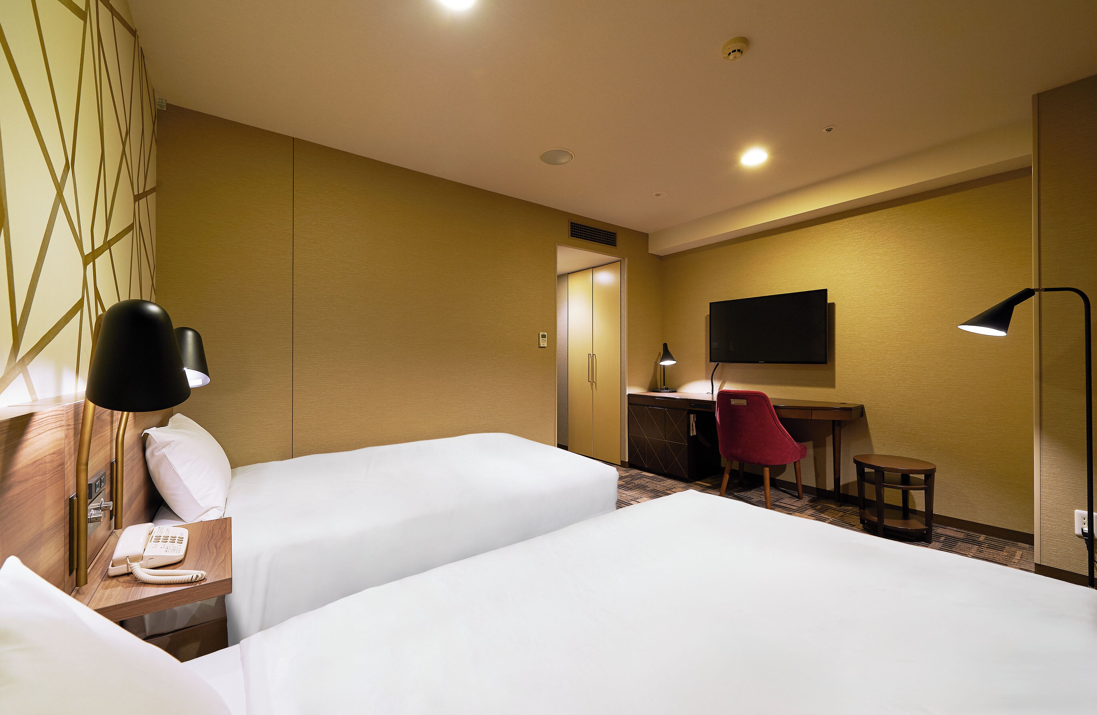 A hotel room with two beds, a desk with a chair, a TV, and a lamp.