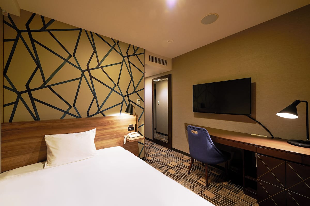 A hotel room with a bed, a desk, a chair, a TV, and a patterned wall.