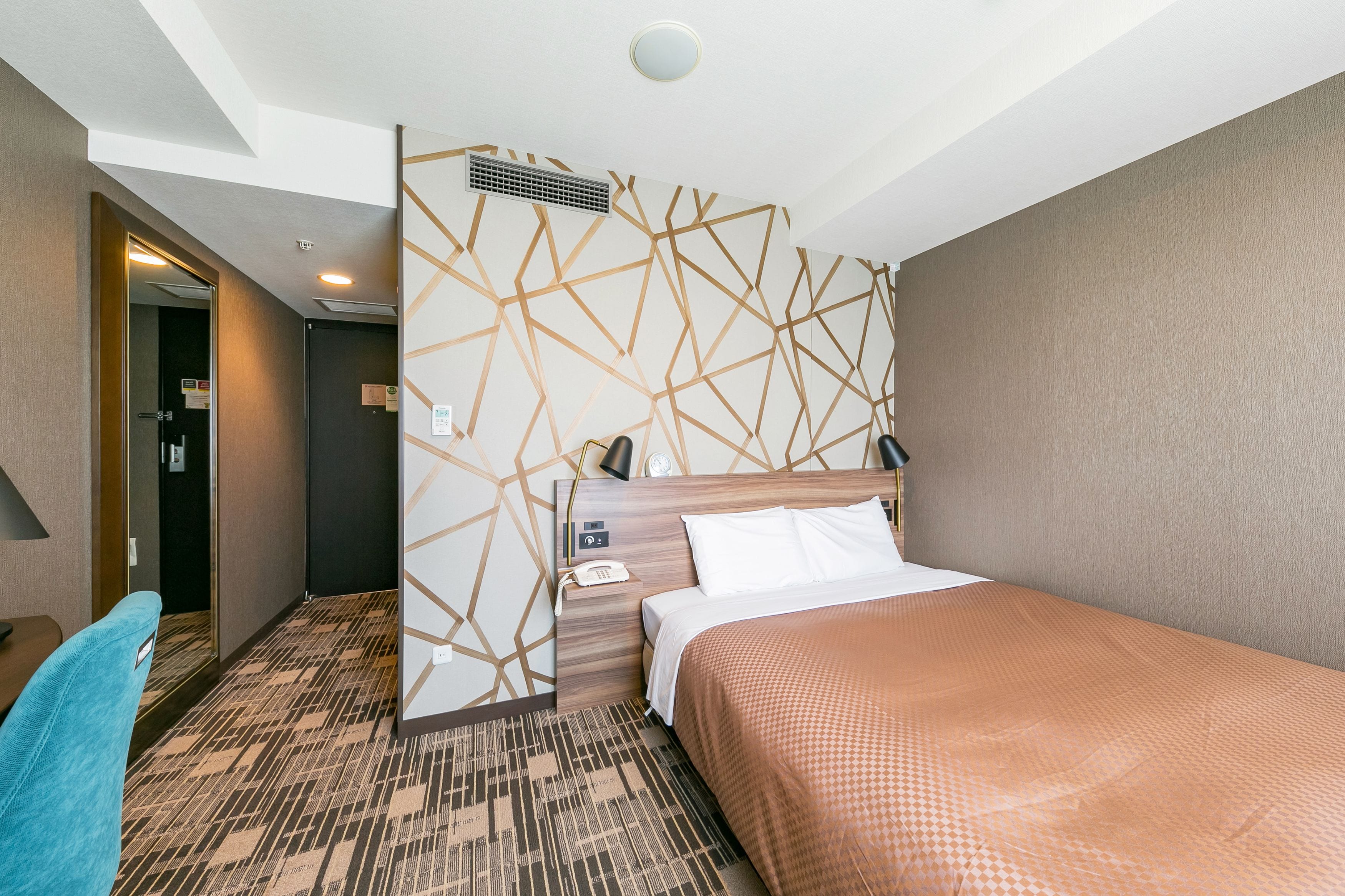A hotel room with a bed, a desk, a chair, a wall with geometric patterns, and a ceiling light.