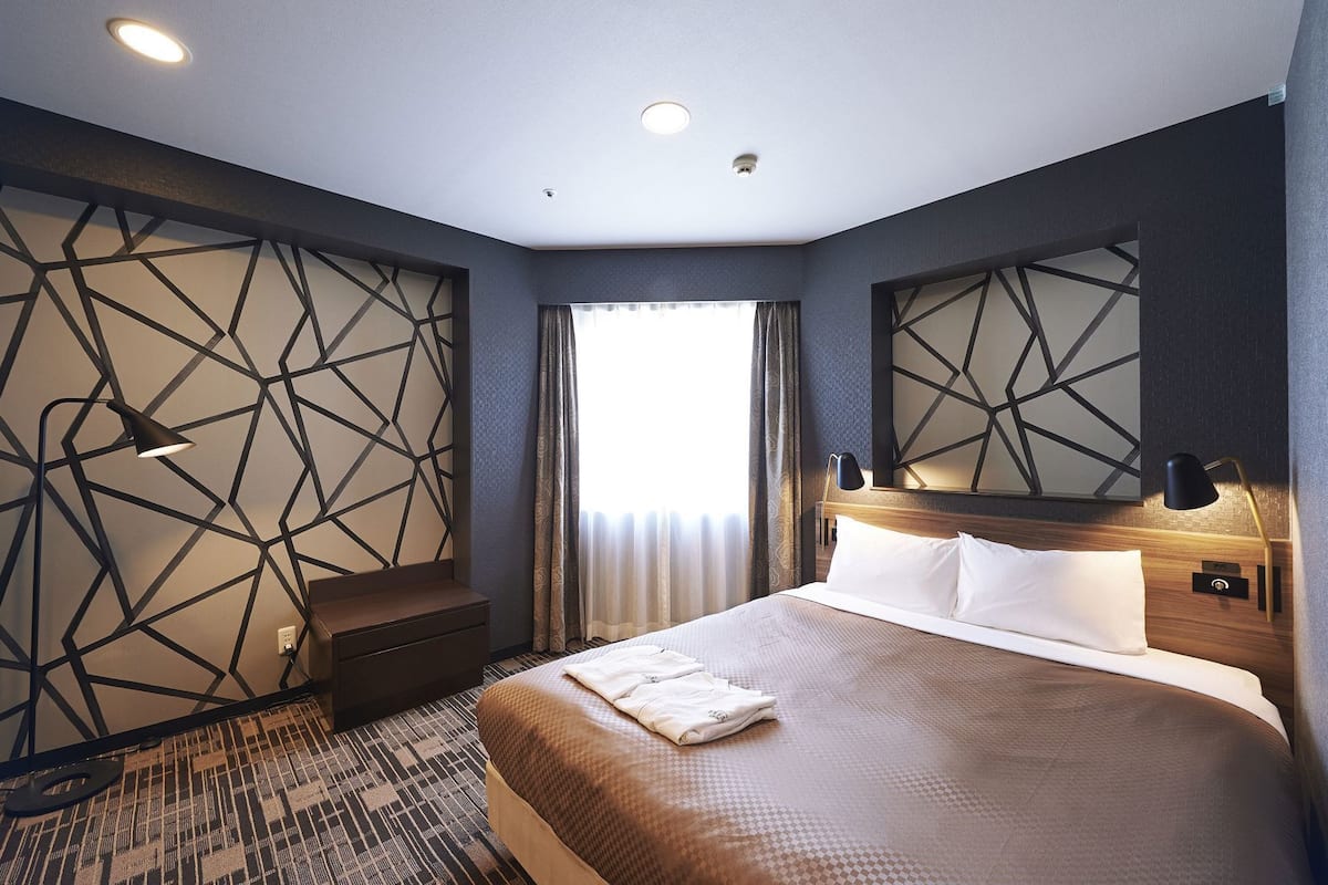 A hotel room with a large bed, a nightstand, a wall-mounted lamp, and a geometric-patterned wall.