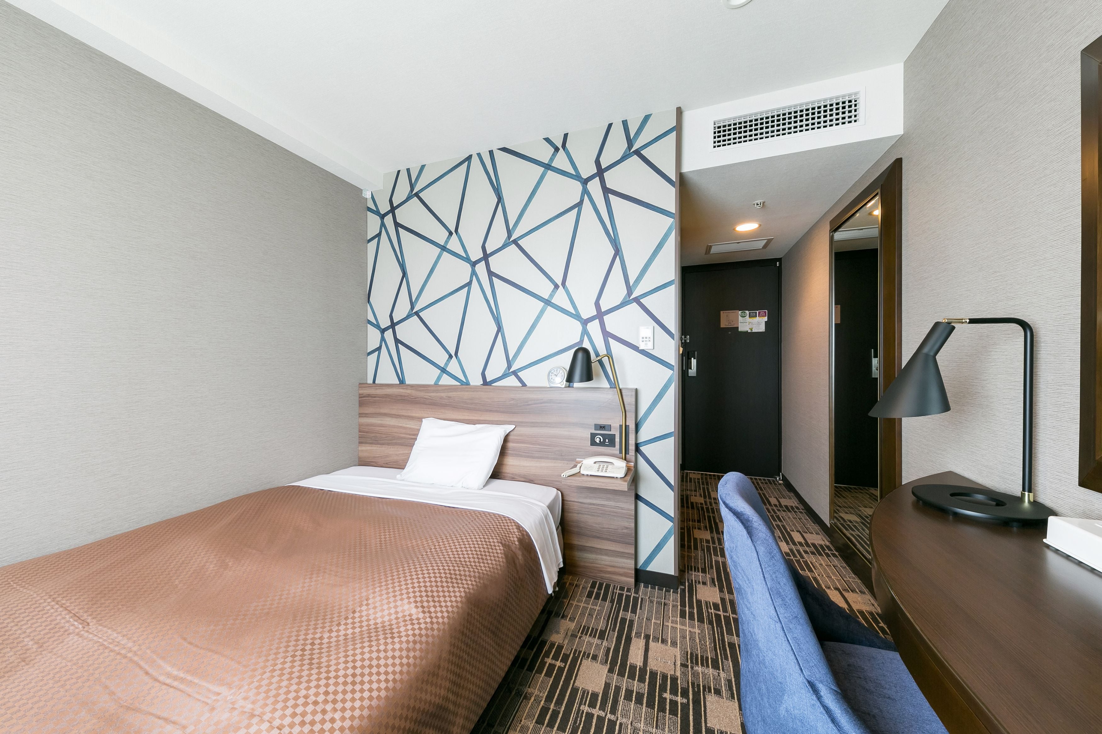 A hotel room with a bed, a bedside table, a lamp, and a patterned wall.
