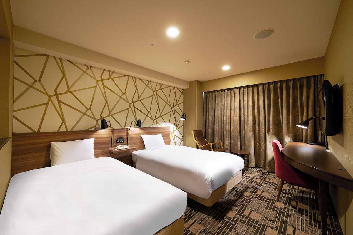 A hotel room with two beds, a desk, a chair, a TV, and a patterned wall.