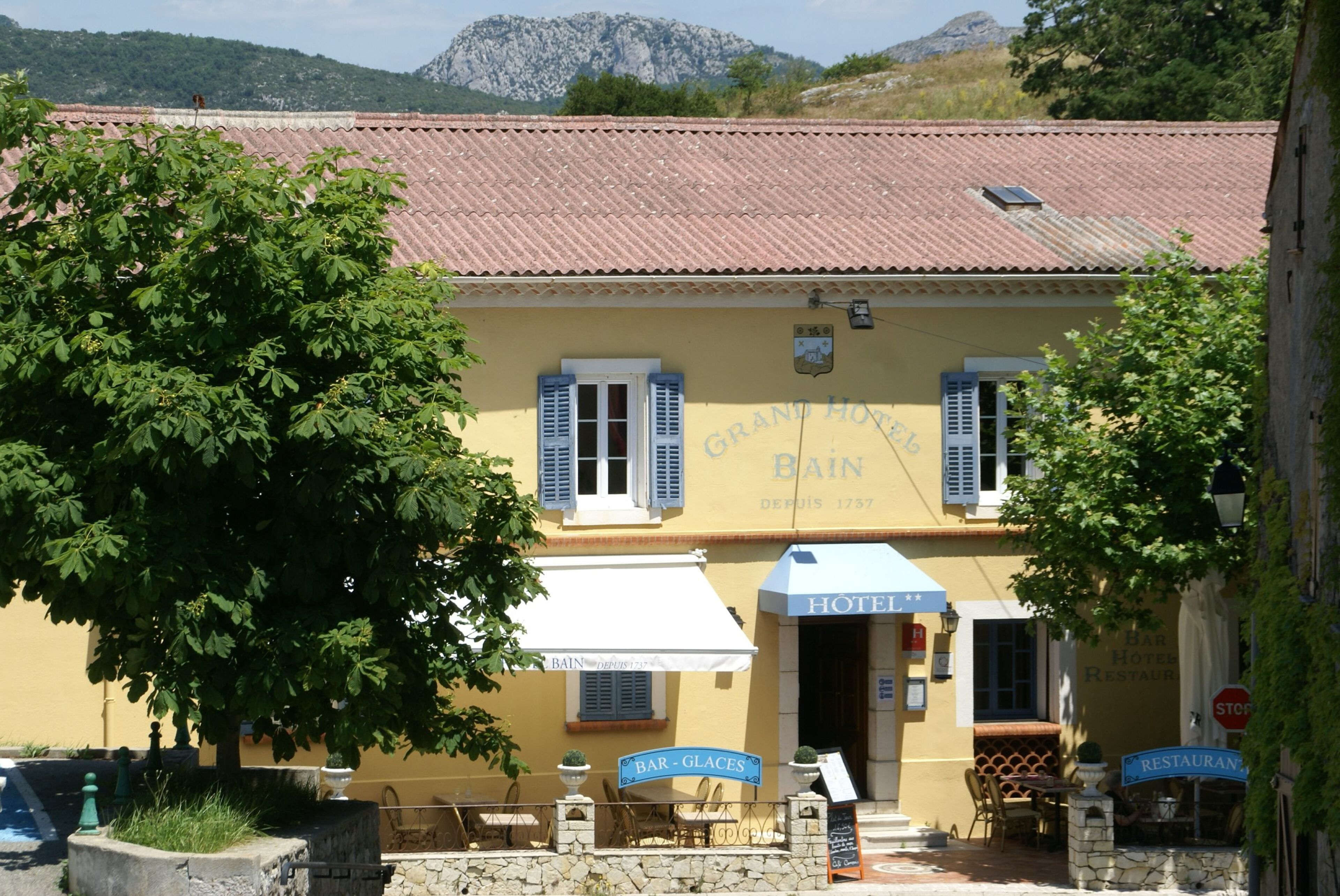 A yellow building with a red-tiled roof, a sign reading "Bain Hotel," outdoor seating, and a mountain in the background.