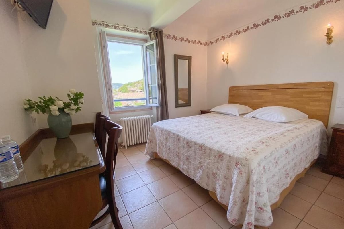 A bedroom with a wooden bed, a desk with a vase of flowers, and a window with curtains.