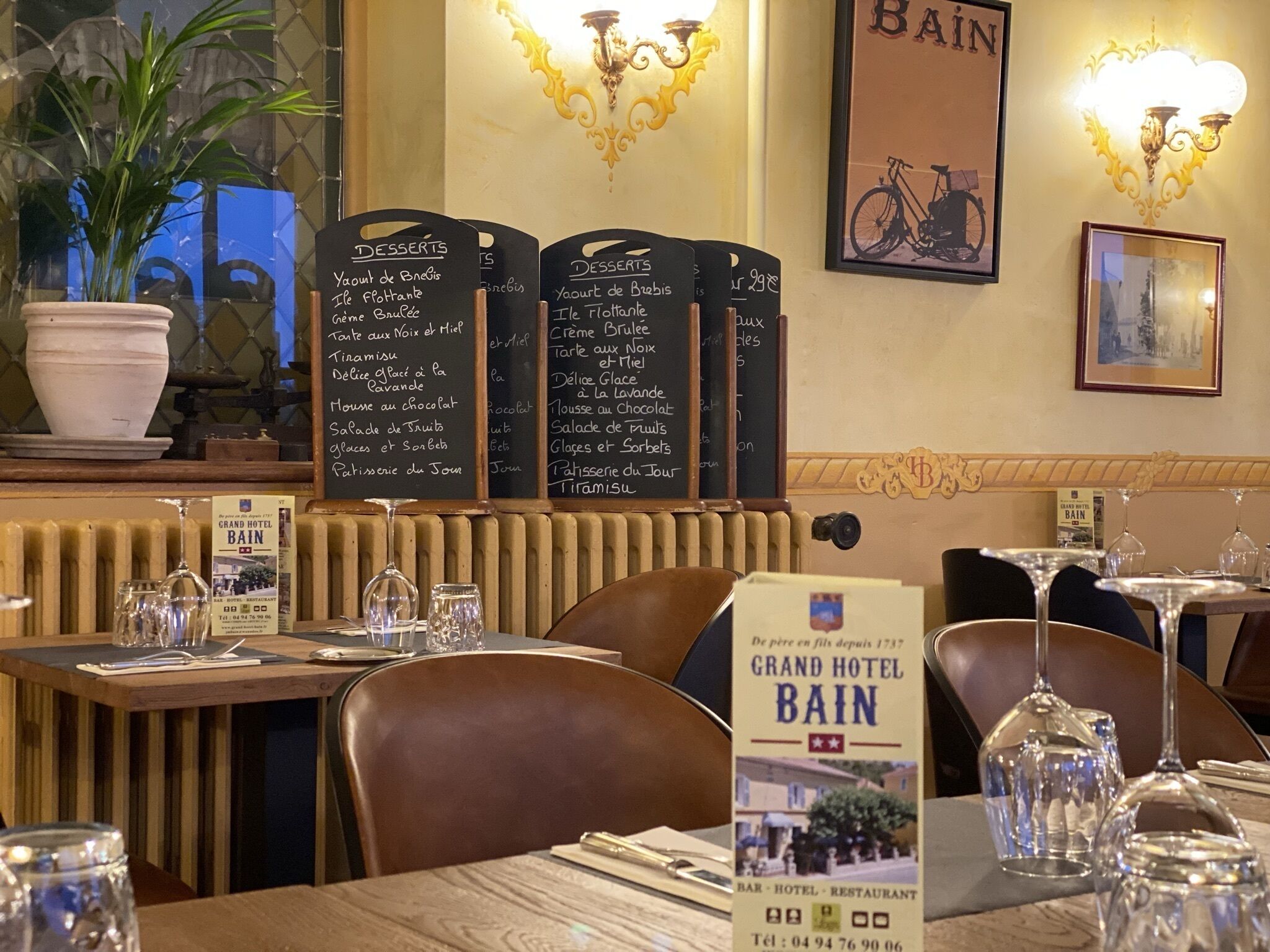 Interior de un restaurante con una pizarra que muestra postres, un letrero del Hotel Grand Bain y una foto enmarcada de una bicicleta en la pared.