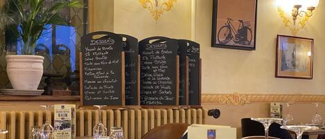 Interior de un restaurante con una pizarra que muestra postres, un letrero del Hotel Grand Bain y una foto enmarcada de una bicicleta en la pared.