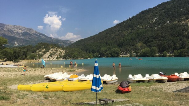 Una playa junto al lago con barcos, una sombrilla azul y gente cerca del agua.