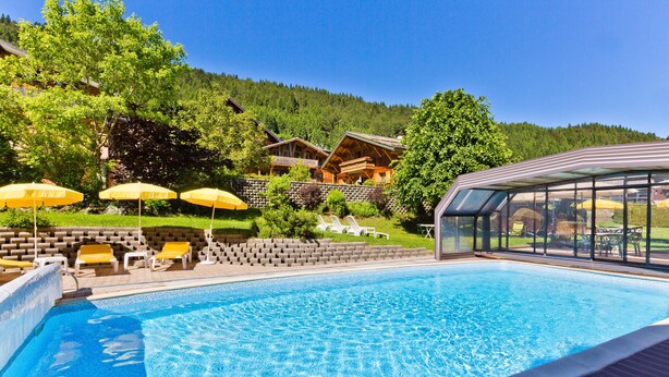 Piscina con sillas de descanso y sombrillas, rodeada por una terraza de madera y un paisaje montañoso.