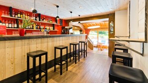 Zona de bar con techo de madera, taburetes, estanterías con botellas y un letrero rojo.
