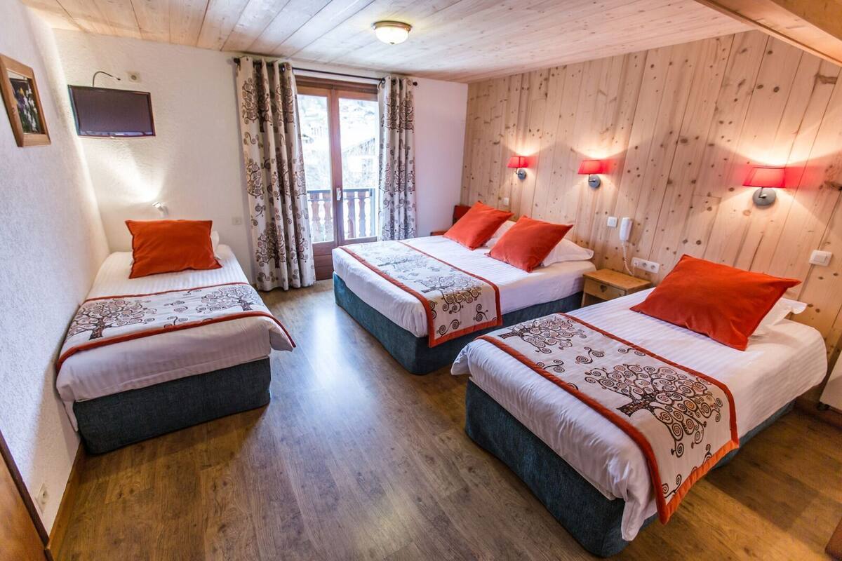 Ein Zimmer mit drei Betten, jedes mit einer gemusterten Tagesdecke und orangefarbenen Kissen. Zwei Wandlampen und ein Fernseher sind an der Wand angebracht.