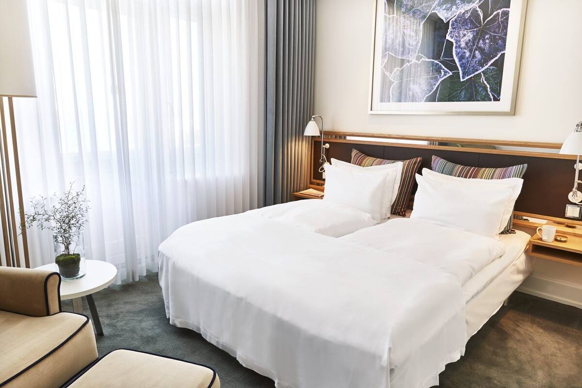 Habitación de hotel con una cama grande, una mesita con una planta, una silla y una obra de arte enmarcada en la pared.