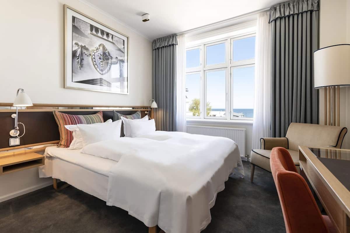 Habitación de hotel con una cama grande, un escritorio, una silla, una lámpara, una ventana con cortinas y un cuadro en la pared.