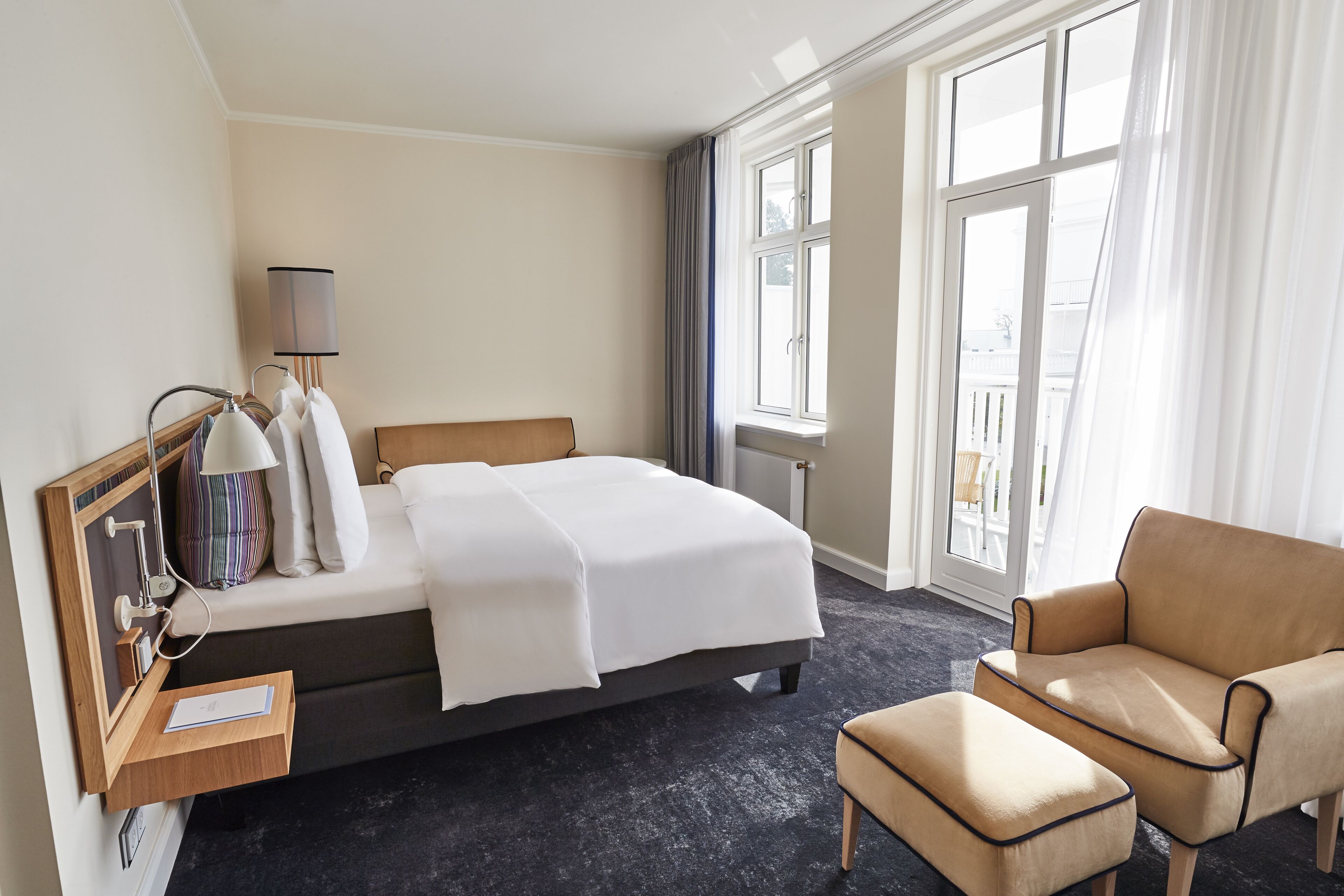 Ett hotellrum med en säng, en stol, ett skrivbord med en lampa och ett fönster med gardiner.