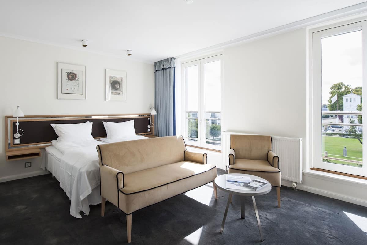 Una habitación de hotel con una cama, un sofá, una silla, una mesita y ventanas amplias.