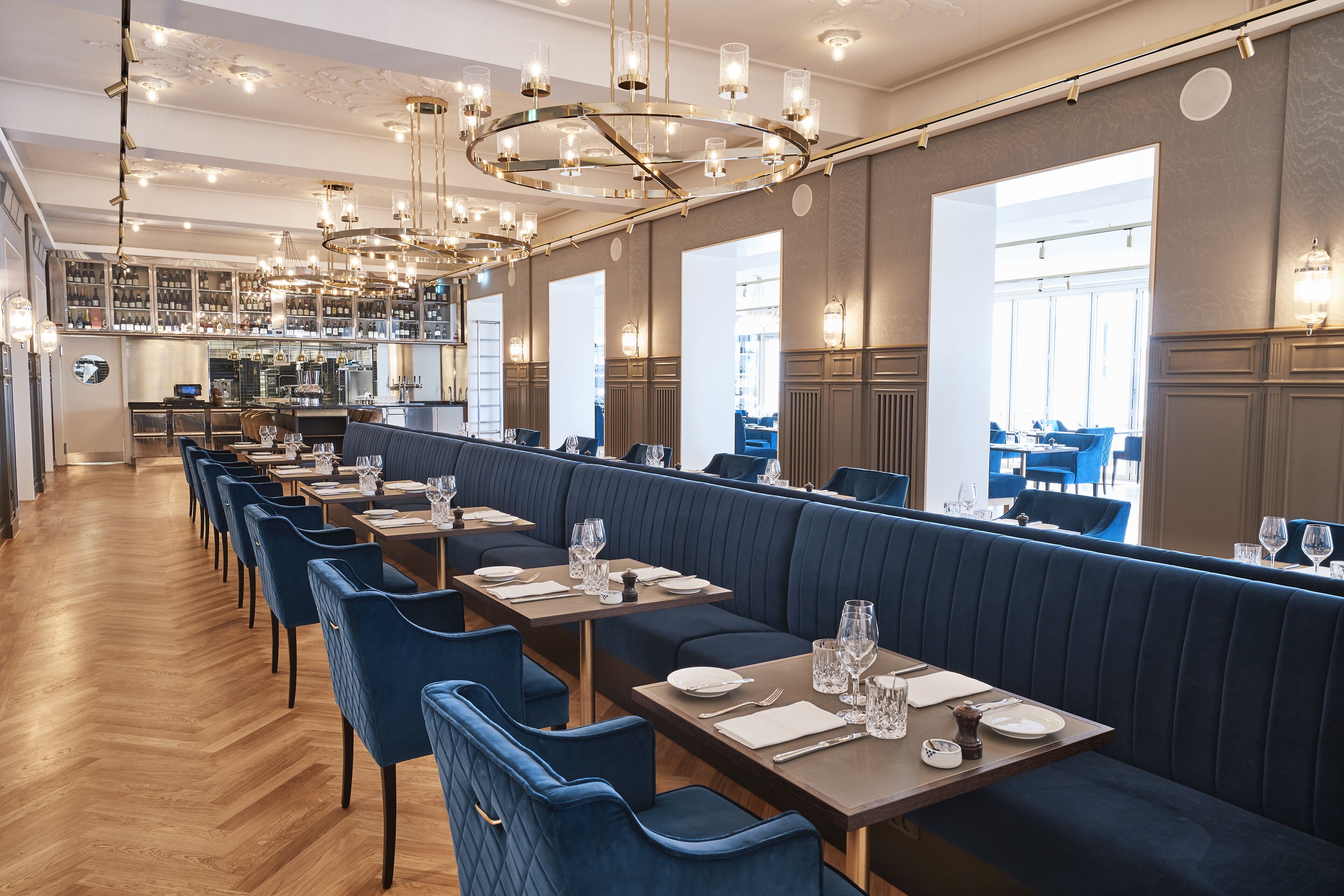 A restaurant with long wooden tables, blue upholstered booths, and a central chandelier.