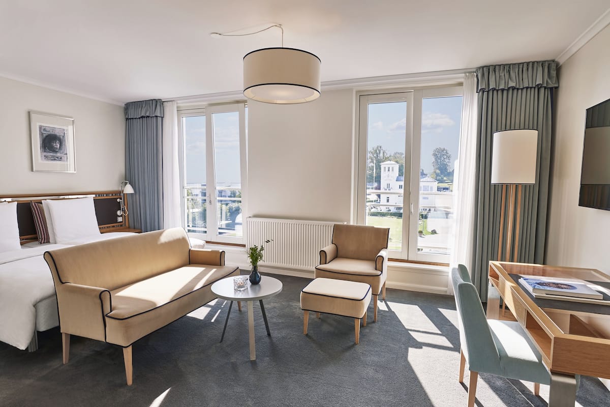 Una habitación de hotel moderna con sofá, sillón, reposapiés y una mesita de noche con lámpara.