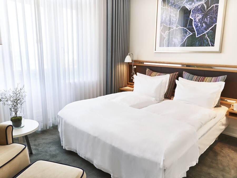 Hotelkamer met een groot bed, twee nachtkastjes, een kleintje met een plantje en een raam met dunne gordijnen.