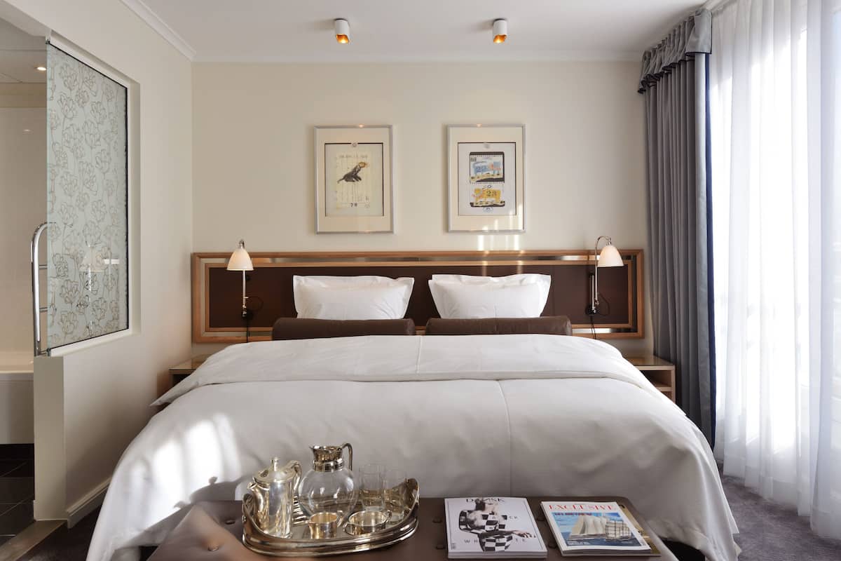 Un cuarto de hotel con una cama grande, dos mesitas de noche con lámparas, una bandeja con vasos y una jarrón, y dos cuadros en la pared.
