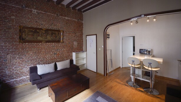 Un salón con pared de ladrillo, un sofá oscuro, una mesa de centro de madera y dos taburetes de bar.