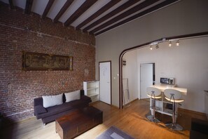 Un salón con pared de ladrillo, un sofá oscuro, una mesa de centro de madera y dos taburetes de bar.