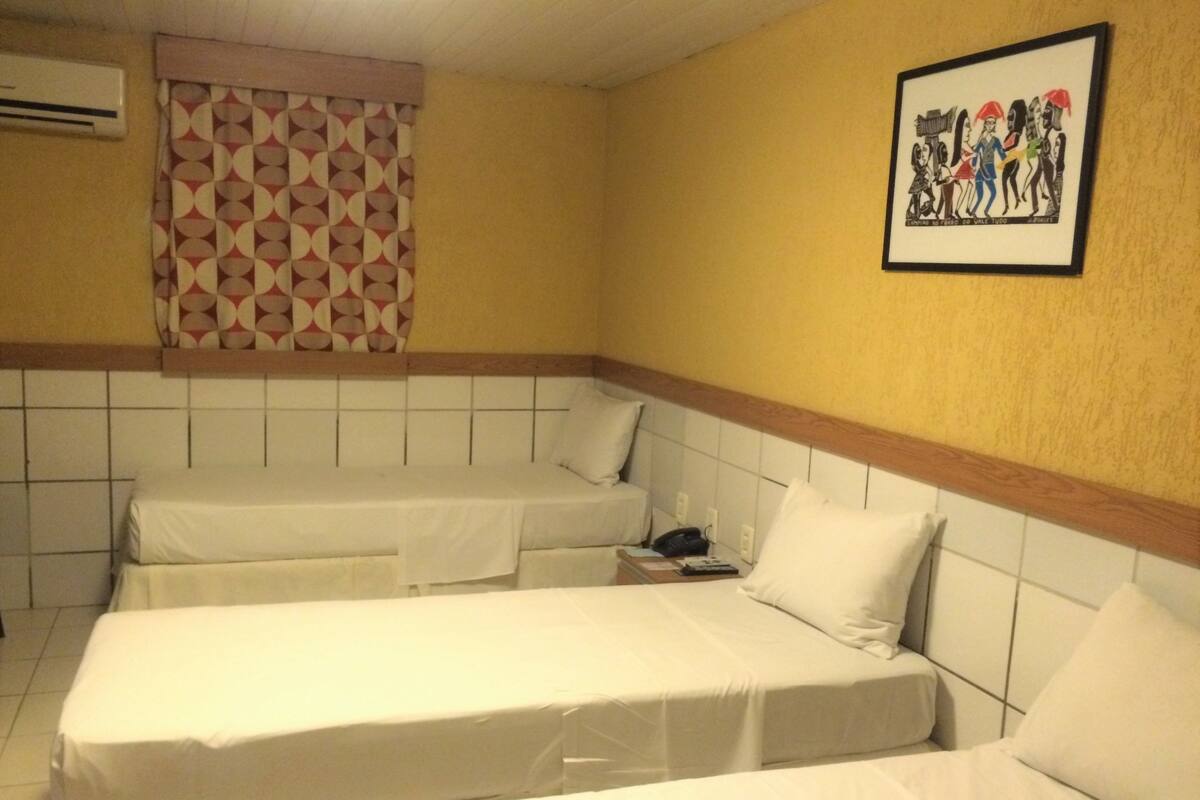 Quarto de hotel com duas camas de solteiro, mesa de cabeceira com telefone e ar condicionado fixado na parede.