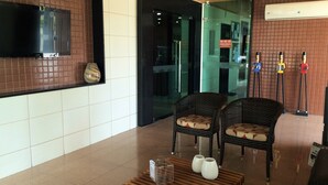 A lobby with a flat-screen TV mounted on a tiled wall, wicker chairs with cushions, a wooden coffee table with vases, and a tiled floor. - Hotel Veraneio (Recife)