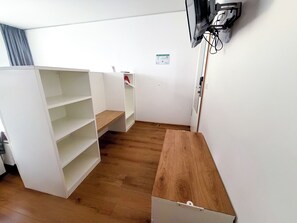 Une pièce aux murs blancs, avec un plancher en bois, un téléviseur fixé au mur et une petite étagère.