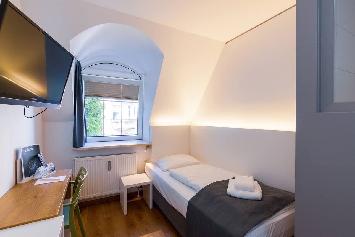A small, well-lit bedroom with a single bed, a desk with a computer, a TV mounted on the wall, and a window with blinds.