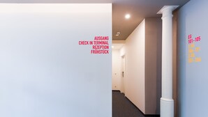 Un pasillo con pared blanca y una columna, con indicaciones de salidas, recepción y registro de entrada.