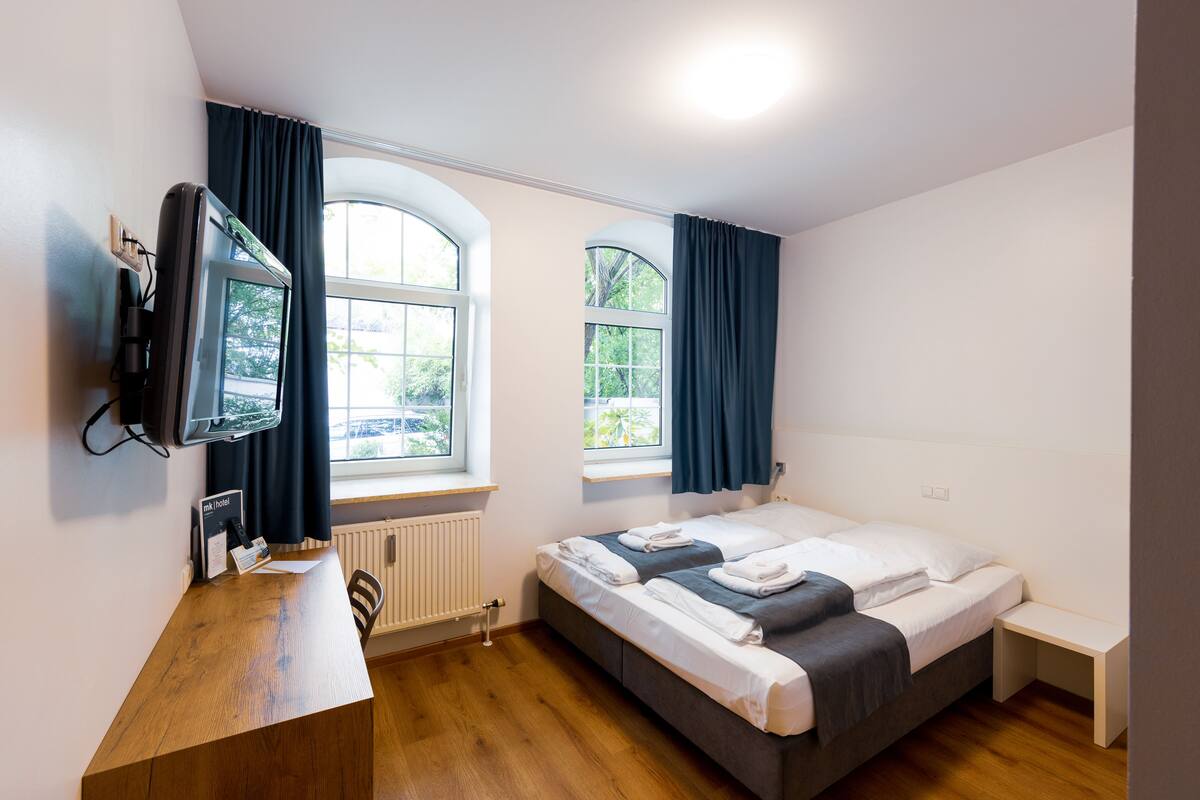 A hotel room with a bed, a desk, a TV, and two windows with curtains.