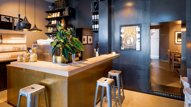 Un área de bar moderna con mostrador de madera, taburetes metálicos y una planta grande en maceta. Hay estantes con botellas y un letrero de neón en la pared.