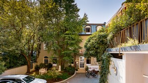 Un hotel con un camino pavimentado, coches aparcados y un jardín con árboles y arbustos.