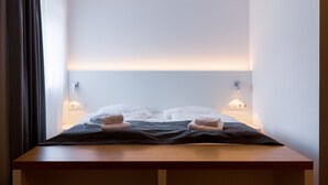 Una cama bien hecha con dos almohadas y dos toallas dobladas, una mesita de noche y una lámpara fijada a la pared.