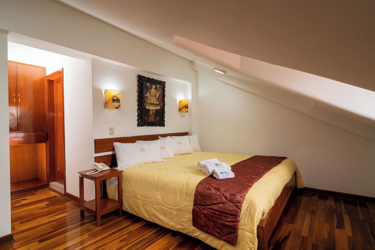 Ein Hotelzimmer mit Holzboden, einem Bett mit beigefarbener Steudecke und roter Tagesdecke, einem Nachttisch aus Holz mit Telefon sowie einem an der Wand hängenden Bild.