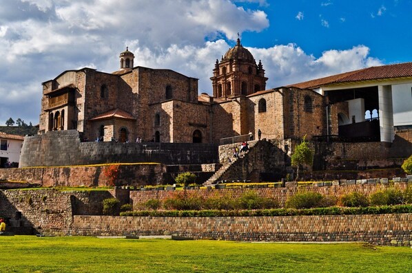 Un edificio histórico de piedra con una imponente torre campanario y una gran cúpula, rodeado de un jardín cuidado y un muro de piedra.