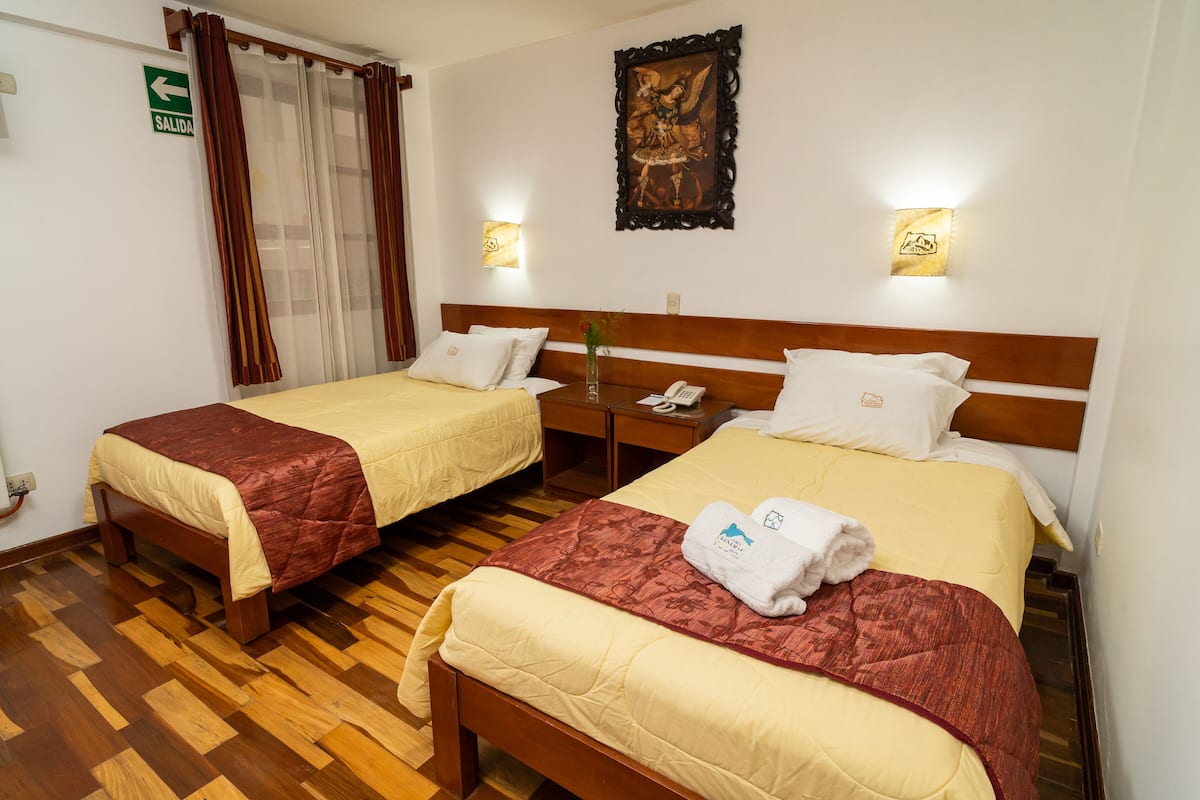 Ein Hotelzimmer mit zwei Betten, Holz-Kopfteilen und Holzboden. An der Wand ist ein grünes Fluchtschild angebracht, über den Betten hängt ein Gemälde und ein Nachttisch steht bereit mit einem Telefon und einer Blumenvase.